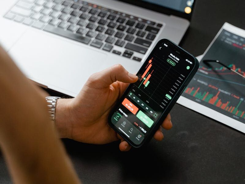 Person analyzing stock market data on a smartphone with a laptop displaying financial graphs in the background.
