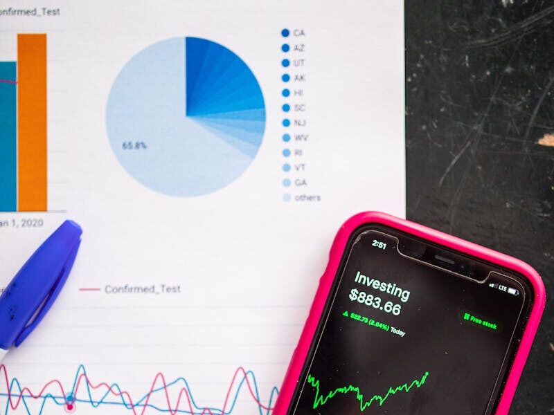 Smartphone displaying stock market data alongside financial charts and a dollar sign note.